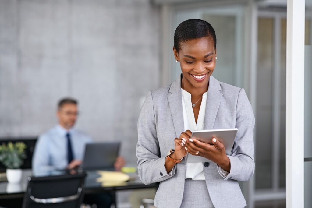 smiling black woman reading tablet with man sat at desk in the background