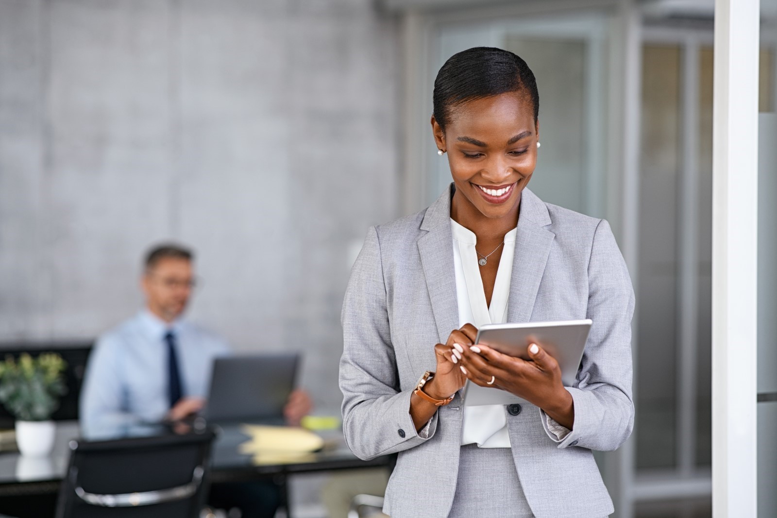 smiling black woman reading tablet with man sat at desk in the background