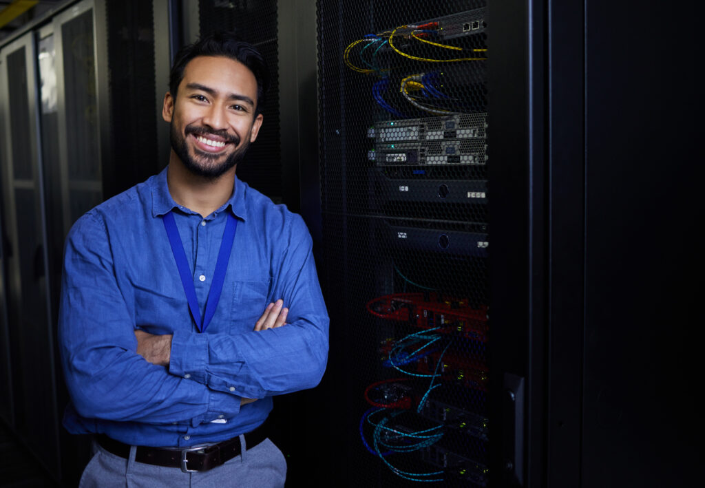 A smiling young man with facial hair stands with crossed arms in front of several dark server racks. The subject wears a blue button down shirt, gray slacks, and a lanyard. Cables and blinking lights are visible on the racks behind him