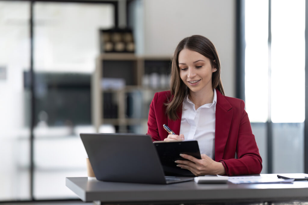 Smiling woman sitting at desk working on laptop taking notes in notebook, happy millennial female working online, watching webinar using computer and writing check list